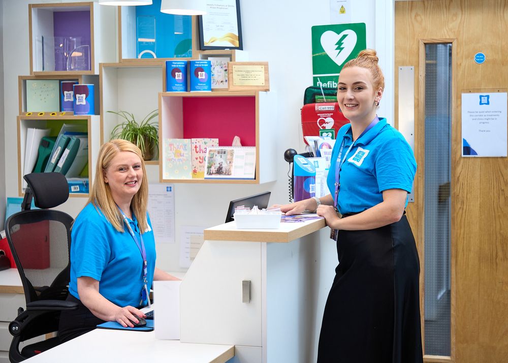Two female receptionists in blue uniforms smile at a modern healthcare reception desk with a defibrillator on the wall.