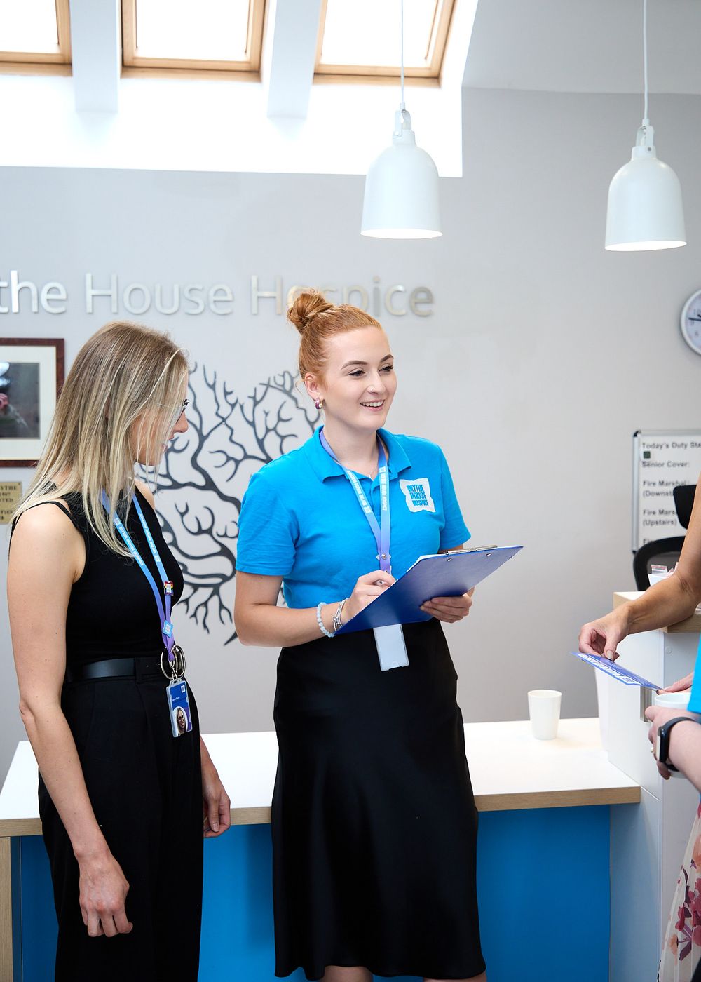 Two hospice staff members in blue uniforms assist a visitor at a reception desk in a bright, welcoming hospice facility.