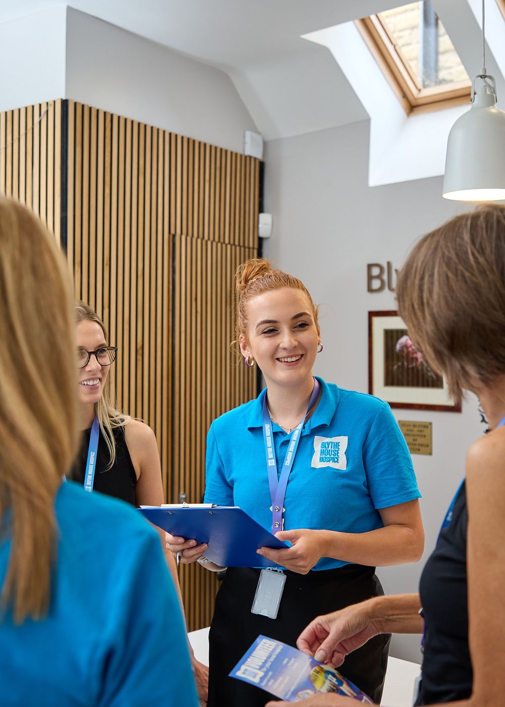 Smiling healthcare worker in blue uniform holds clipboard while discussing with colleagues in a bright modern reception area.