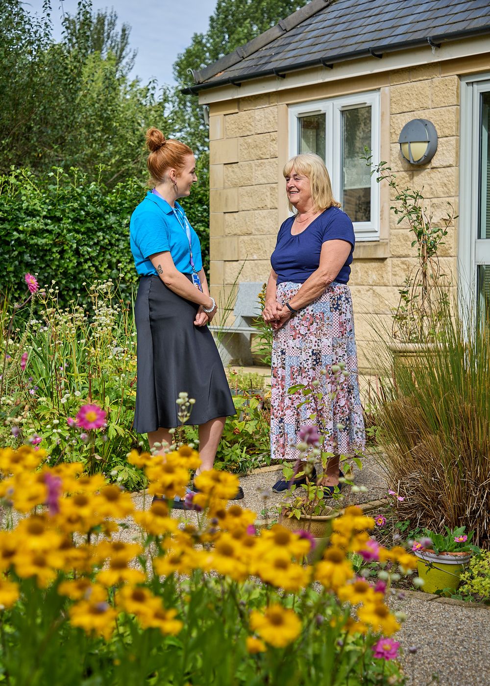 Two women conversing in a sunny cottage garden surrounded by colorful wildflowers in bloom.