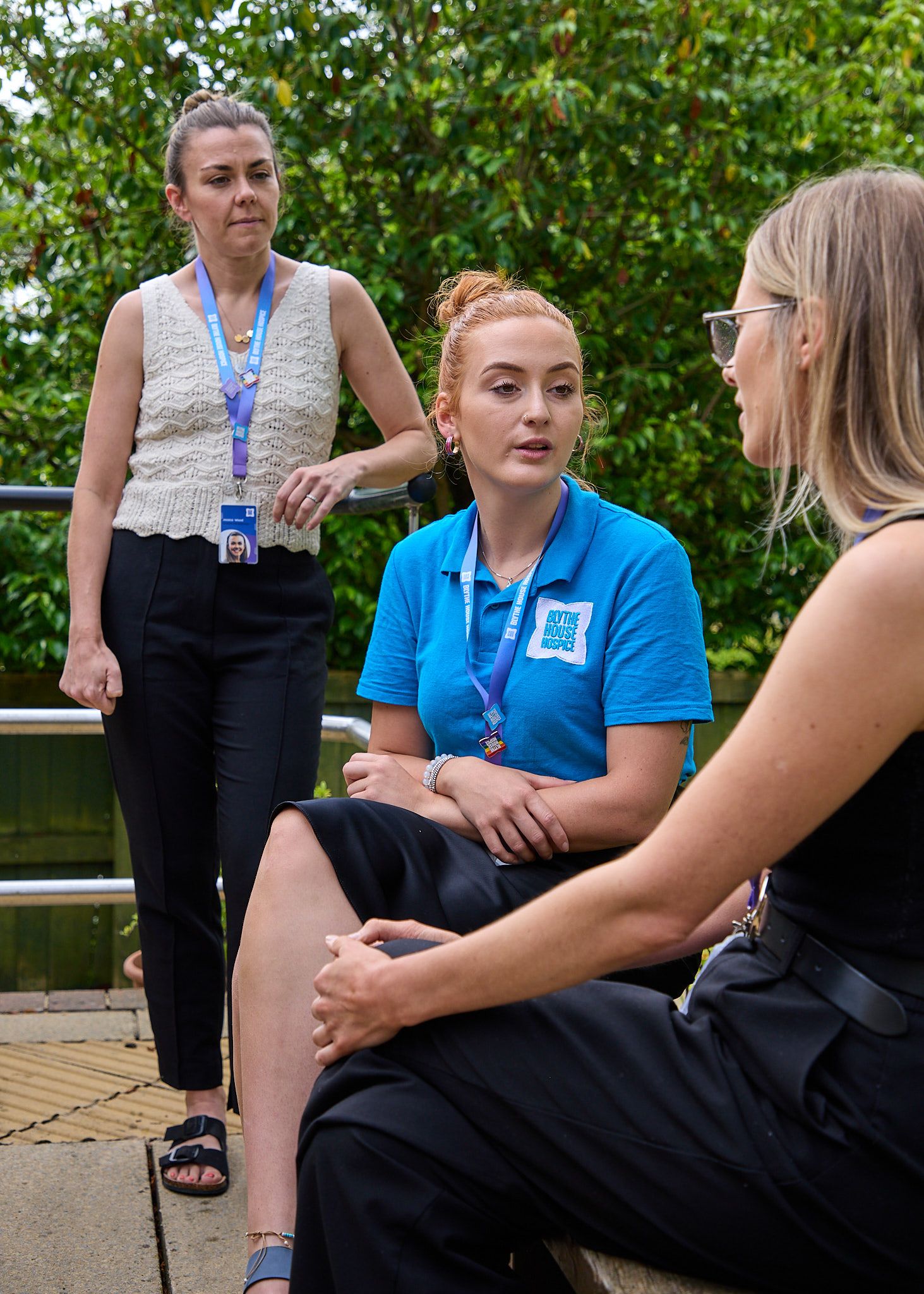 Three women having an outdoor conversation, with one wearing a blue polo shirt and lanyard in a garden setting.