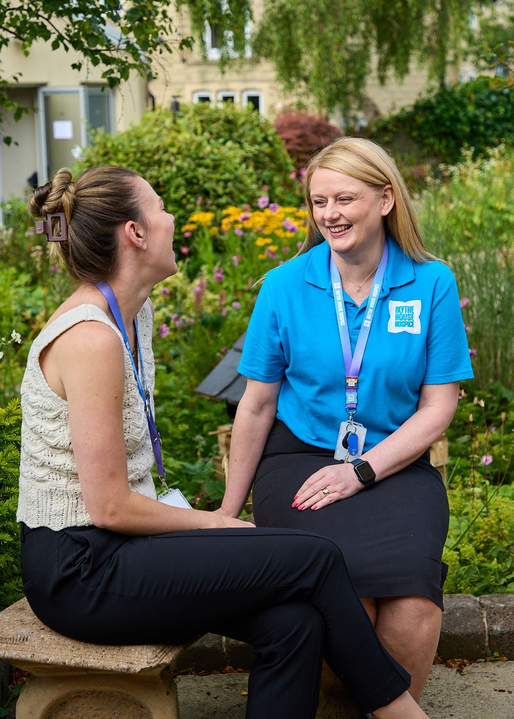 Two women sitting outdoors in a garden, one wearing a blue polo shirt with a lanyard, smiling.