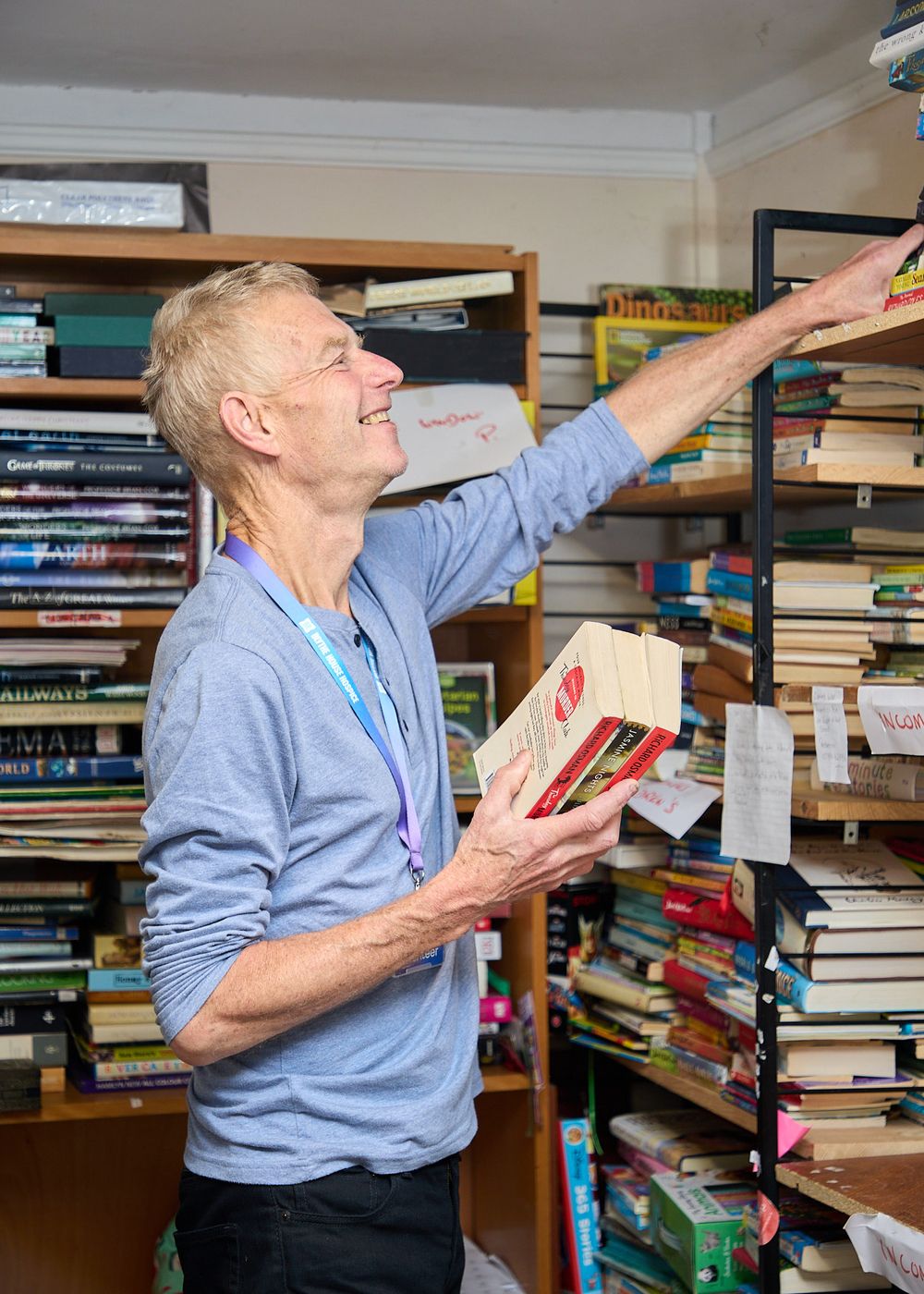 Tall man in blue shirt reaching for books on high shelves in a cluttered secondhand bookshop.