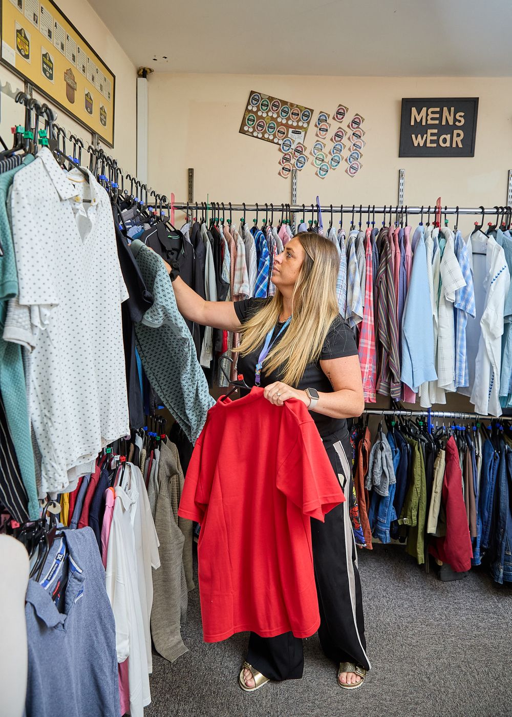 Woman browsing red clothing on rack in charity shop with mens wear section visible in background.