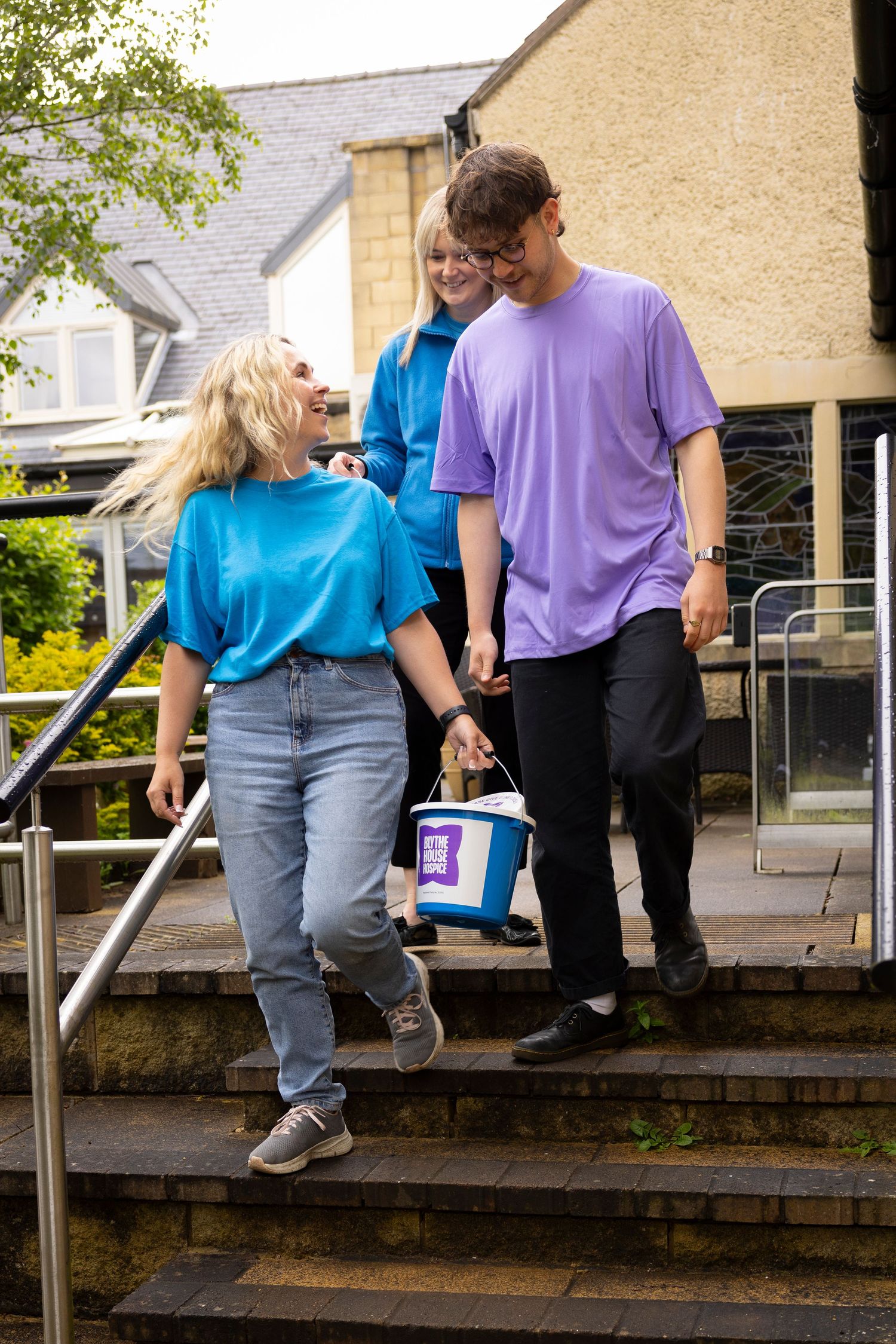 Three young volunteers in blue and purple t-shirts walking down outdoor steps carrying a charity collection bucket.