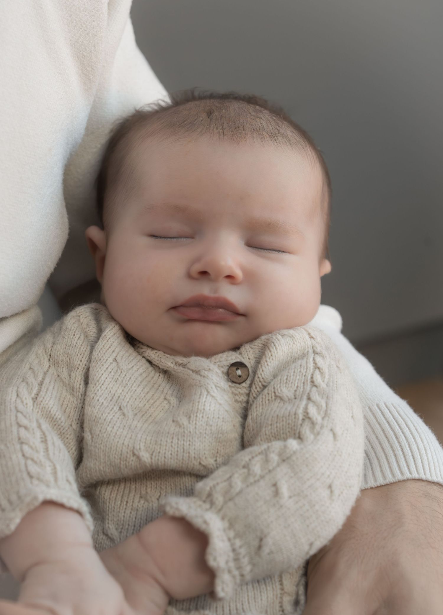 Peaceful newborn baby sleeping in a knit cream cable-knit onesie, cradled in adult arms.