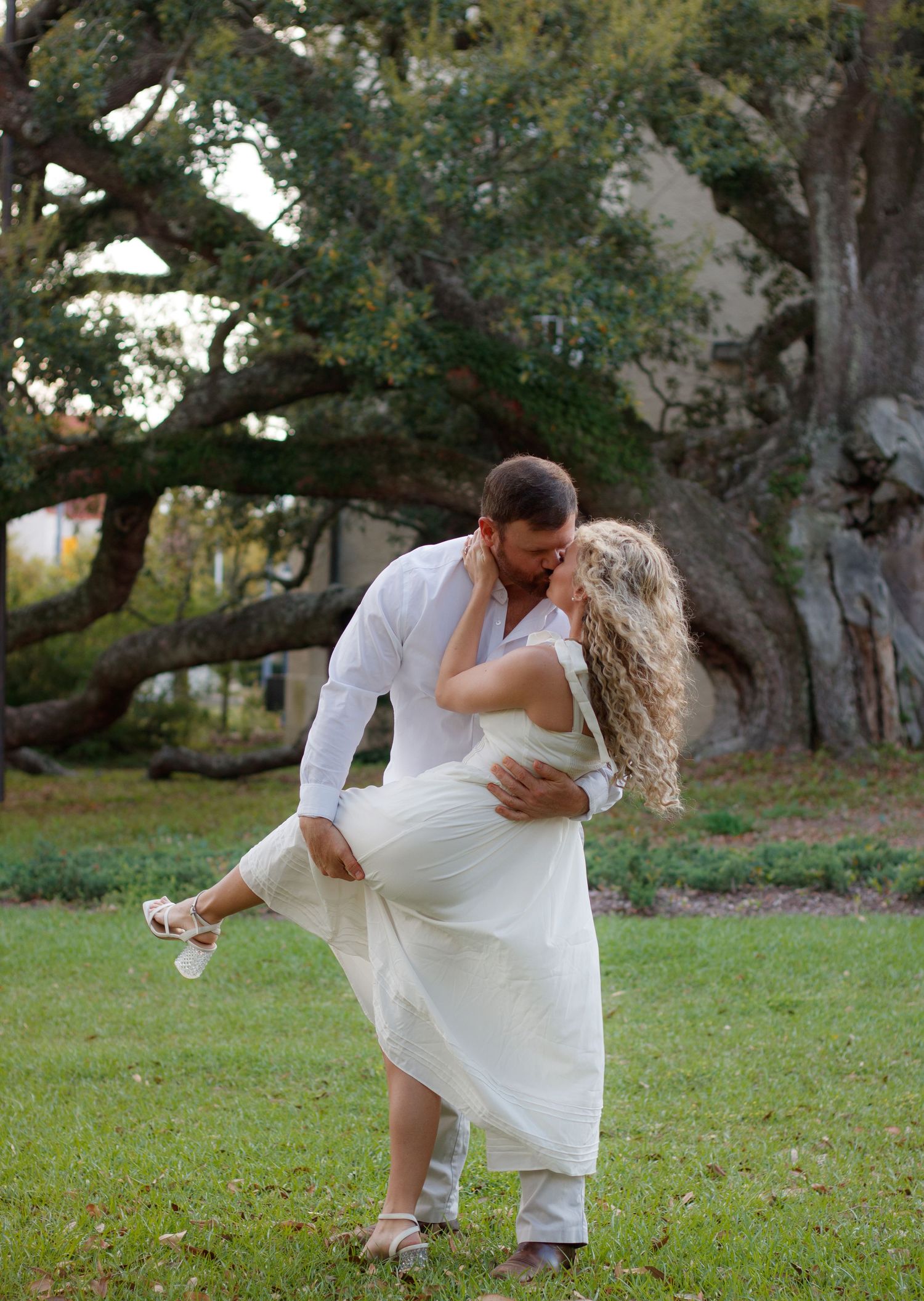 Groom in white shirt dipping and kissing his bride in a flowing cream dress on a grassy lawn beneath a large oak tree.
