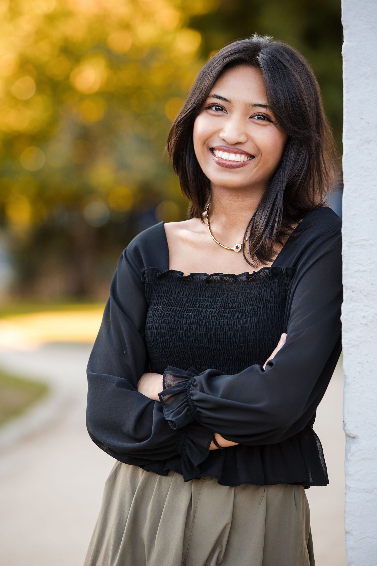 Smiling young woman in black smocked top and olive skirt leaning against white pillar outdoors in autumn.