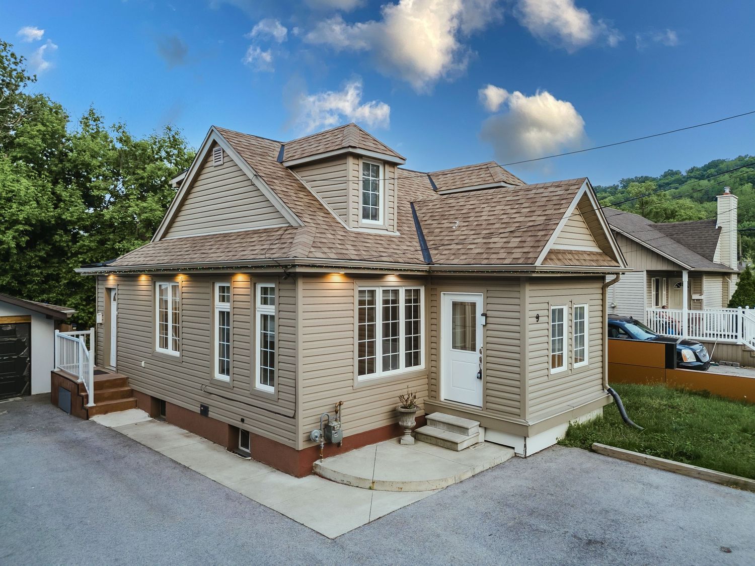 Charming tan vinyl-sided cottage-style home with dormer windows, covered entry, and manicured landscaping on a concrete driveway.