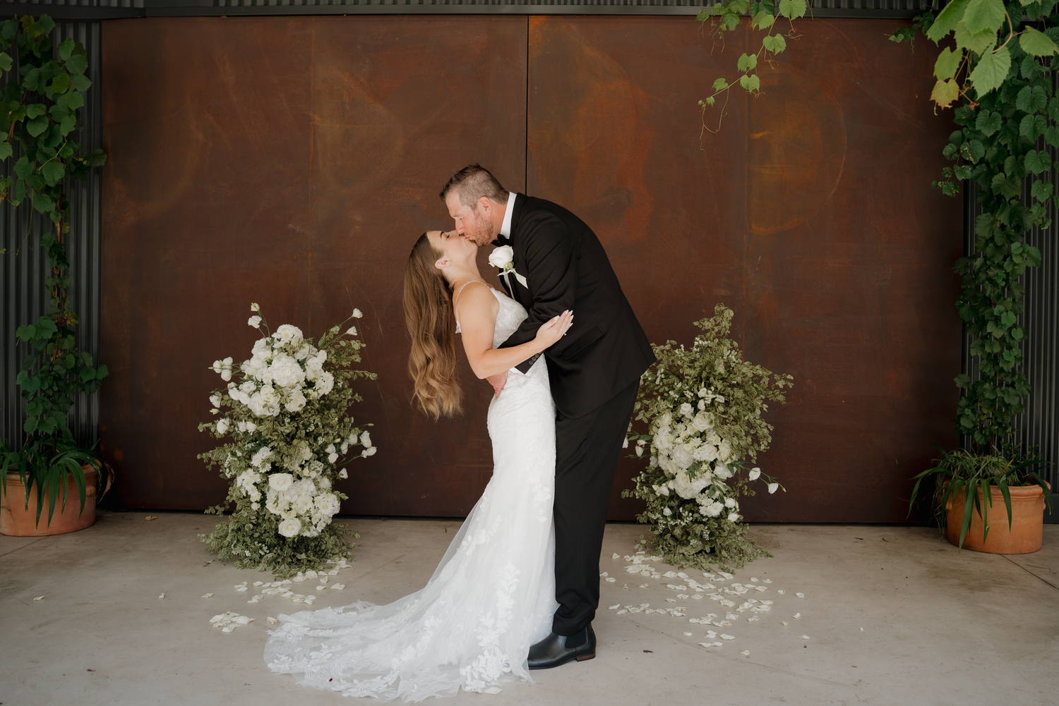 Bride and groom share first kiss at elegant wedding ceremony with white floral arrangements and lush greenery backdrop.