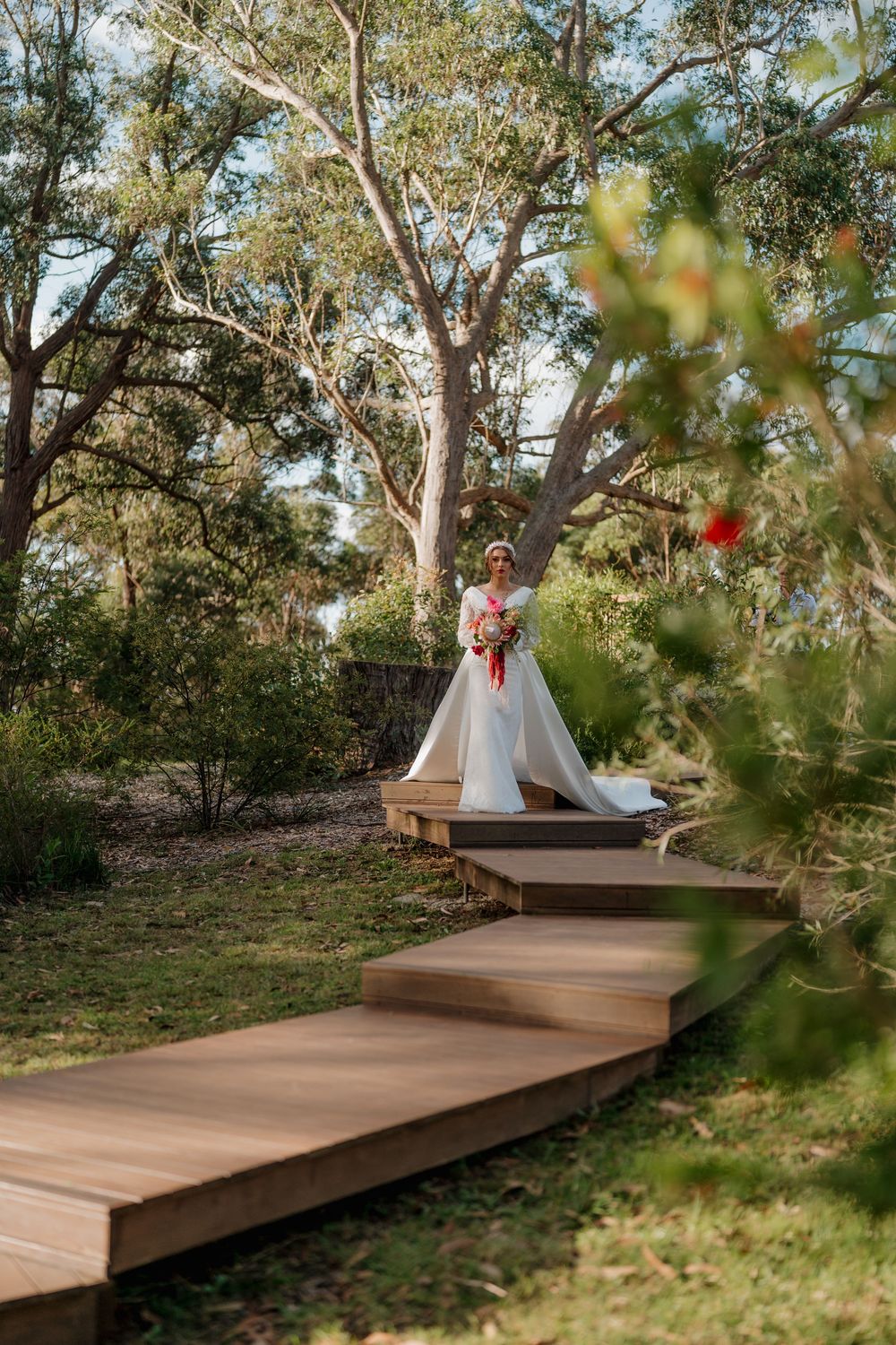 Bride in white gown holding bouquet stands on wooden steps surrounded by lush Australian bushland trees.
