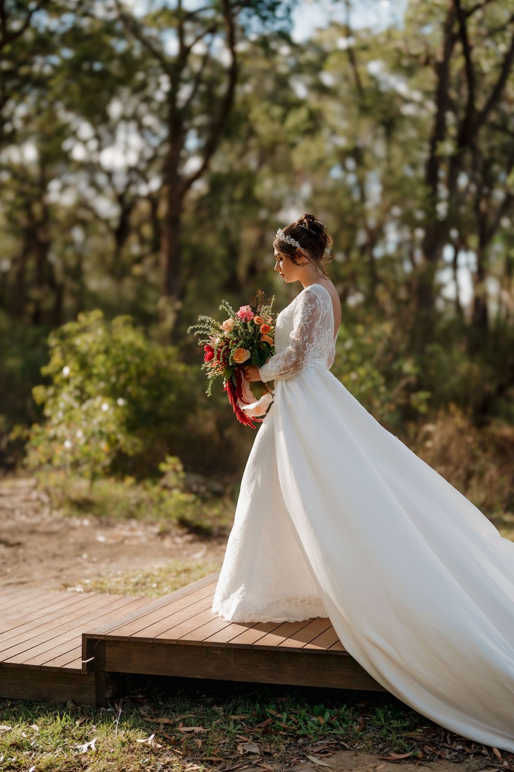 Bride in lace wedding gown holds vibrant bouquet on wooden deck surrounded by forest.