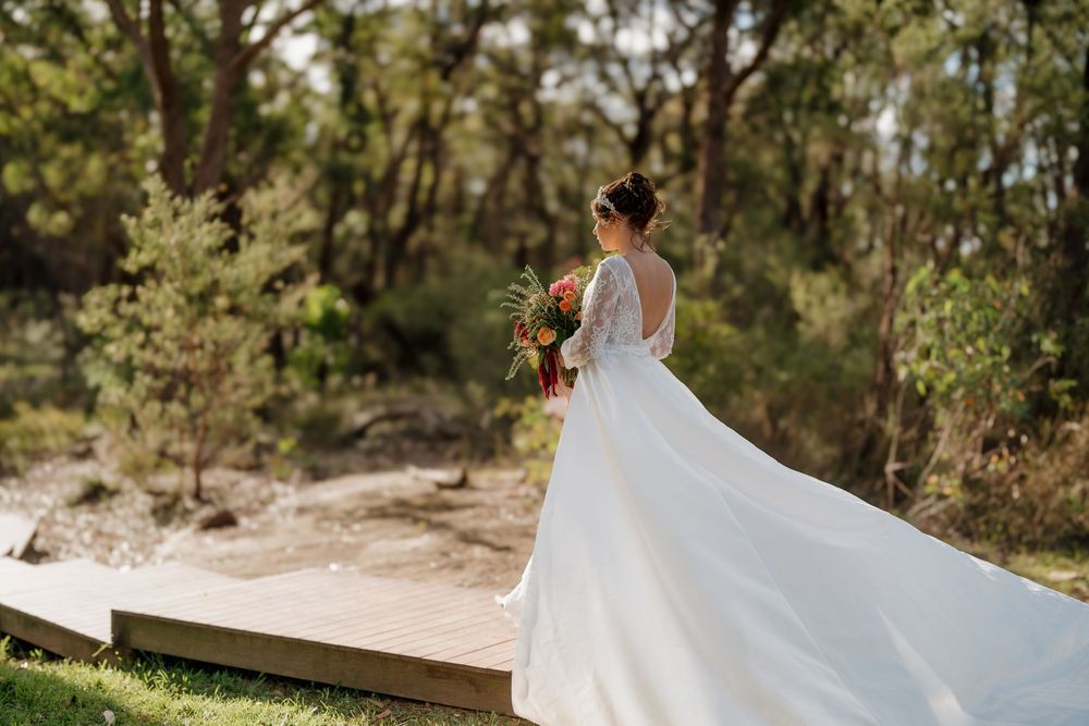 Elegant bride in flowing white gown with long train holding colorful bouquet in a sunlit outdoor setting.