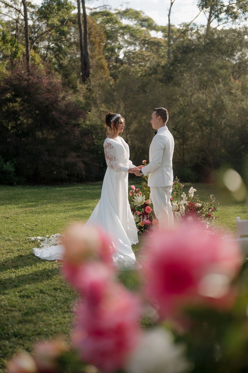 Bride and groom exchanging vows outdoors in a lush garden setting surrounded by pink floral arrangements.