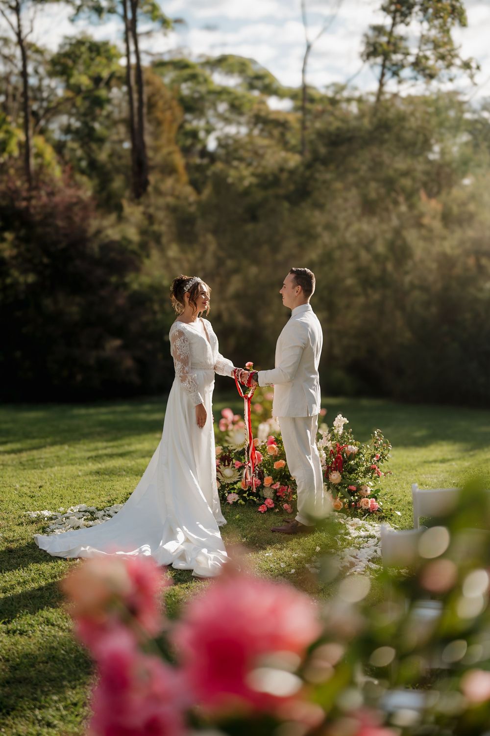 Bride and groom hold hands during outdoor wedding ceremony surrounded by lush greenery and pink floral arrangements.