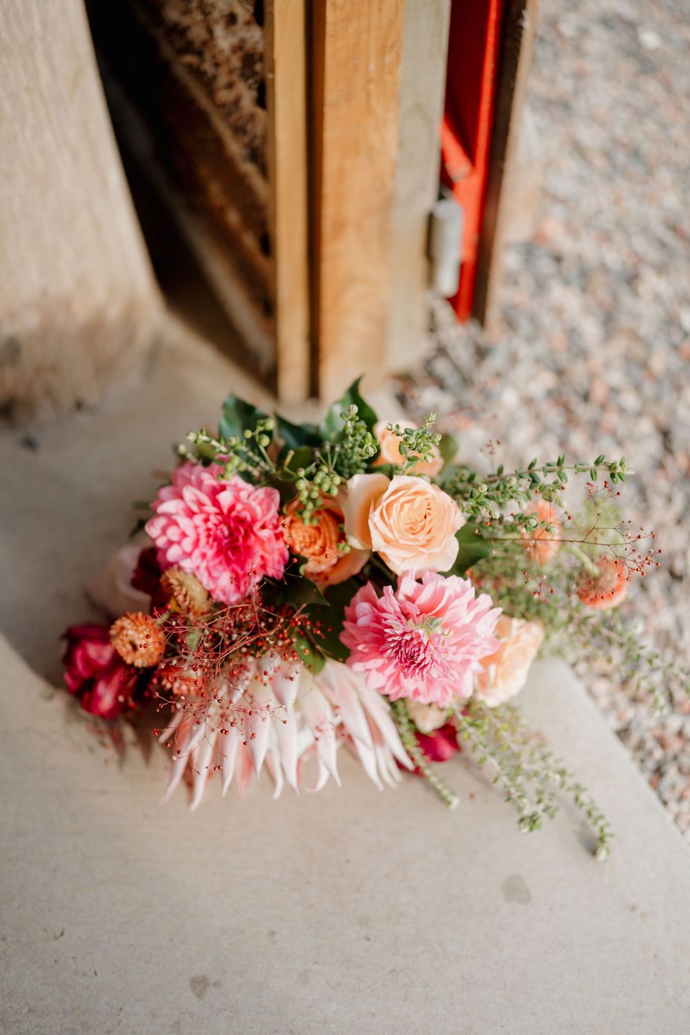 A vibrant wedding bouquet with pink dahlias, peach roses, and greenery resting on a concrete step.