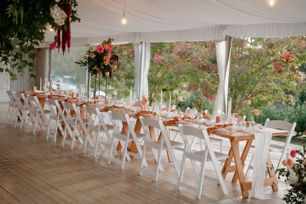 Elegant outdoor wedding reception tent with long tables, white folding chairs, floral centerpieces, and string lights.