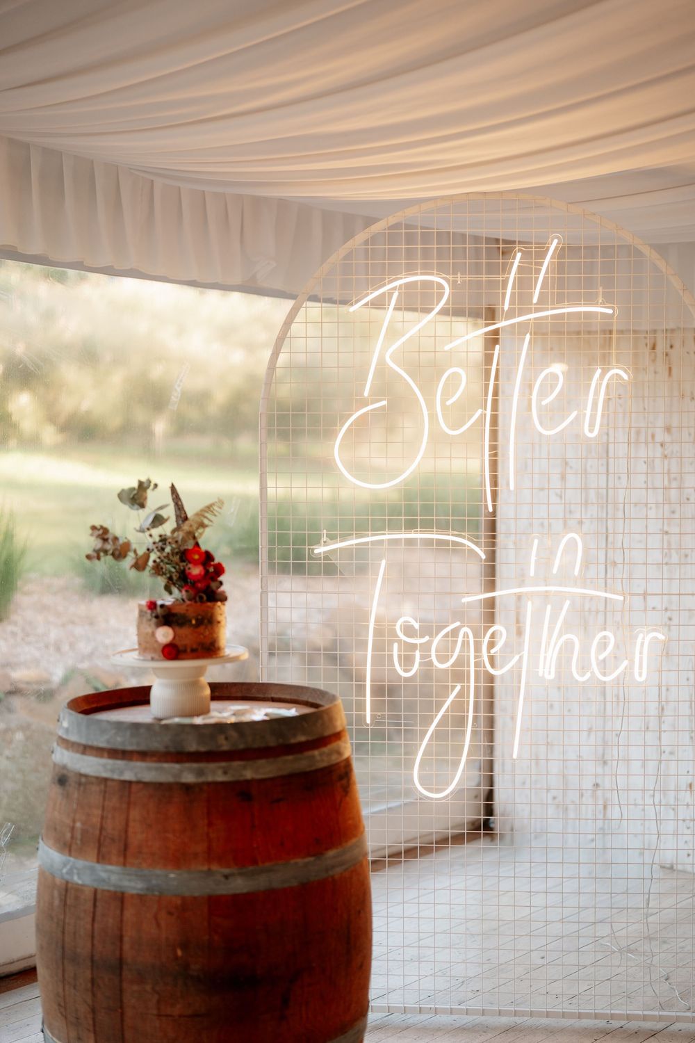 Rustic wedding cake on a wooden barrel with a Better Together neon sign in a draped tent venue.