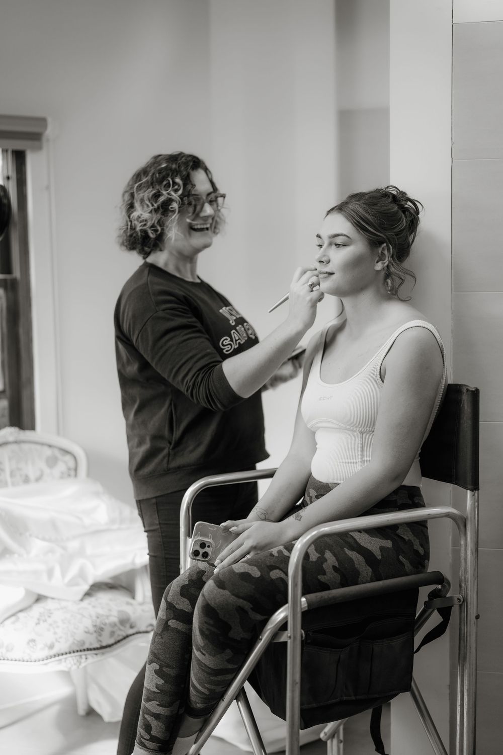 Black and white photo of a makeup artist applying makeup to a bride seated in a wheelchair before her wedding.