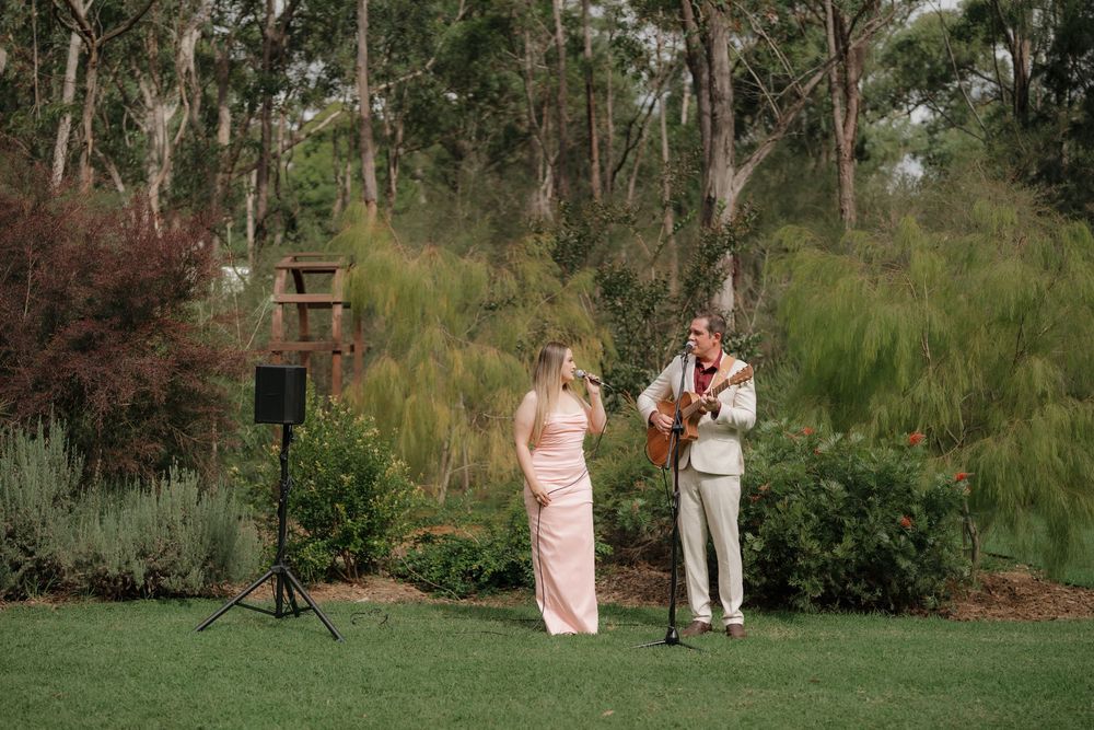 A woman in a pink gown and a guitarist perform outdoors at a garden wedding surrounded by lush green trees.