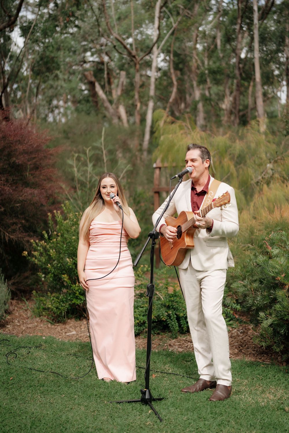 A woman in a pink gown and man in a beige suit perform outdoors, singing and playing guitar at a garden wedding.