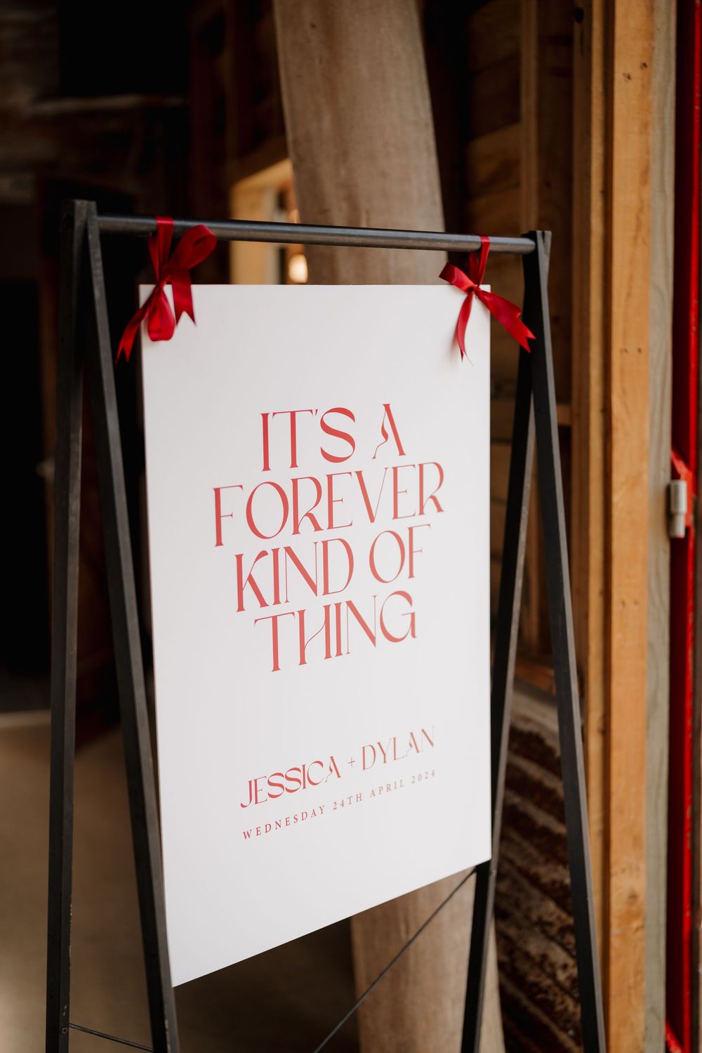 White wedding welcome sign reading It's A Forever Kind Of Thing for Jessica and Dylan, adorned with red ribbon bows.