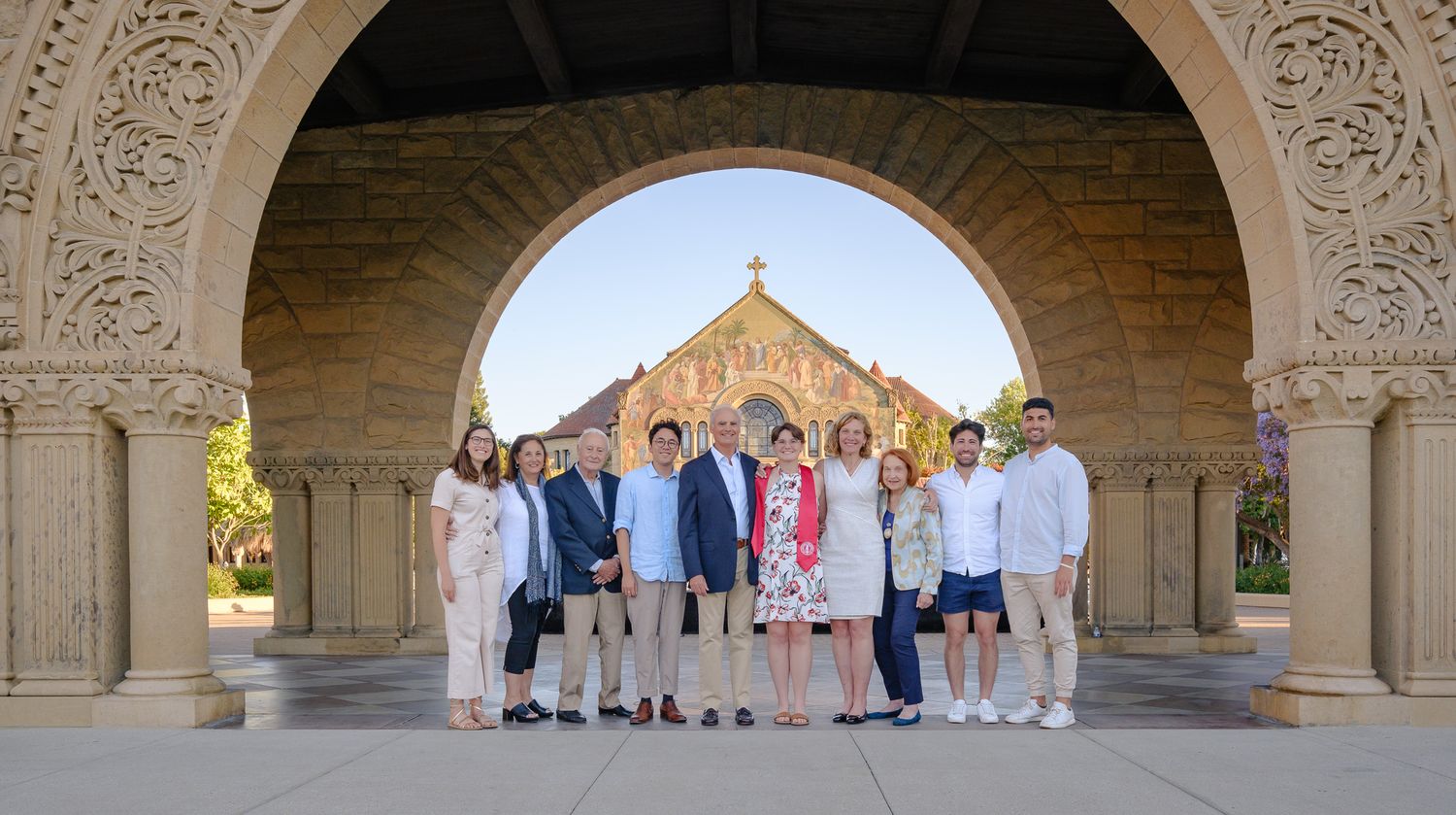 Group of people posing under ornate arch at Stanford University with Memorial Church visible in background.