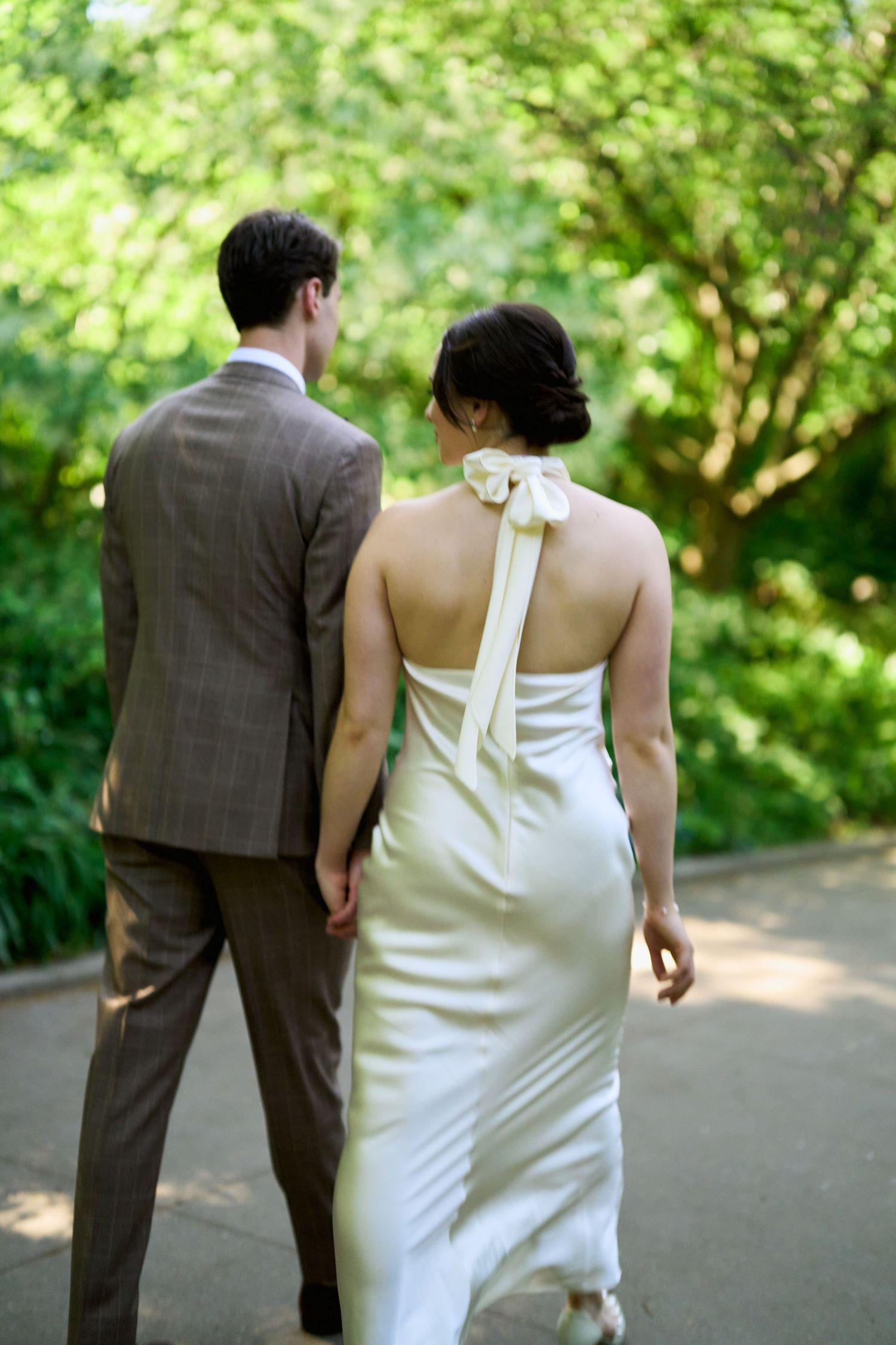 Bride and groom walking hand in hand through a lush green park, bride wearing elegant white halter gown with bow.