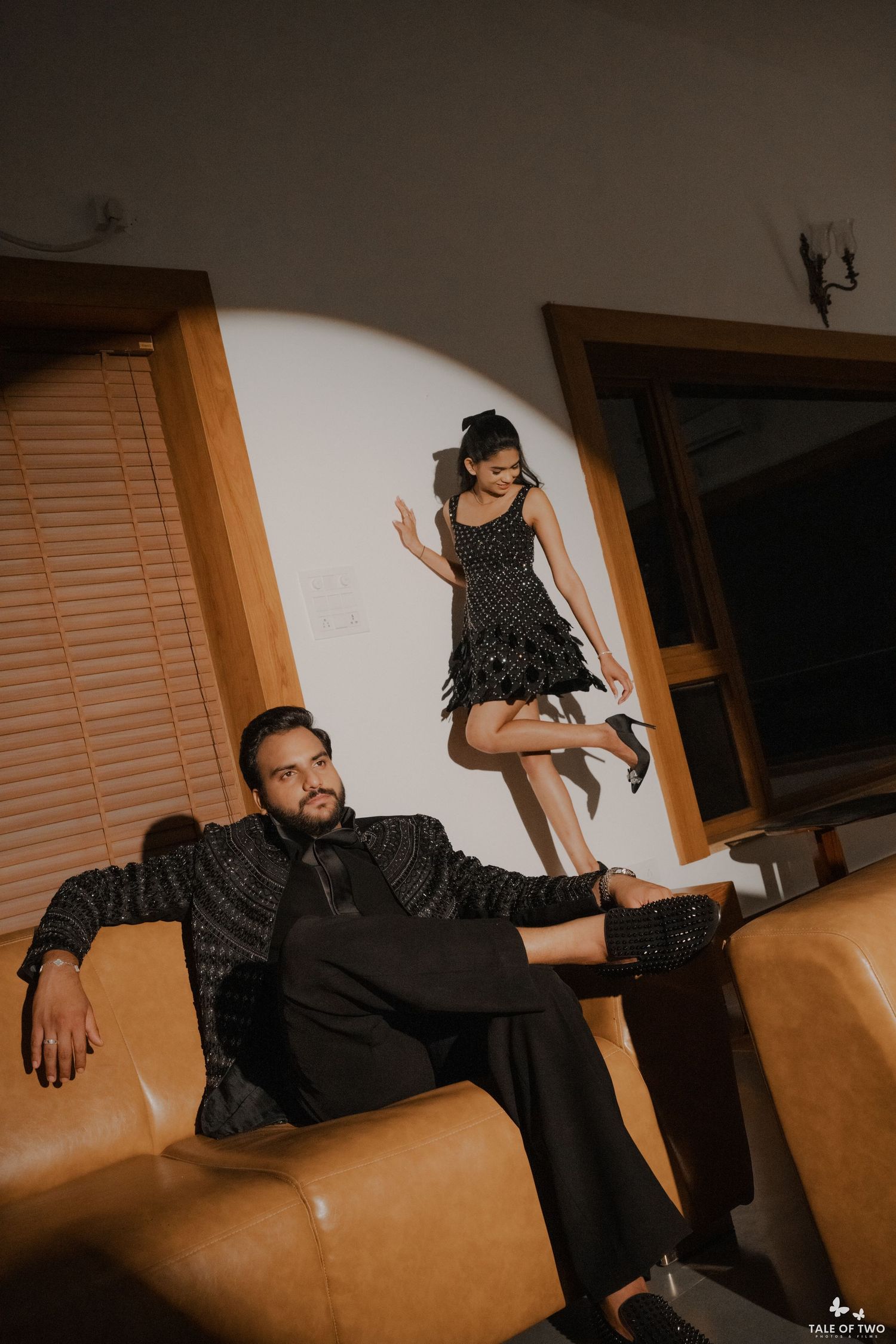Couple in matching black outfits posing elegantly in a dimly lit room with leather sofa.