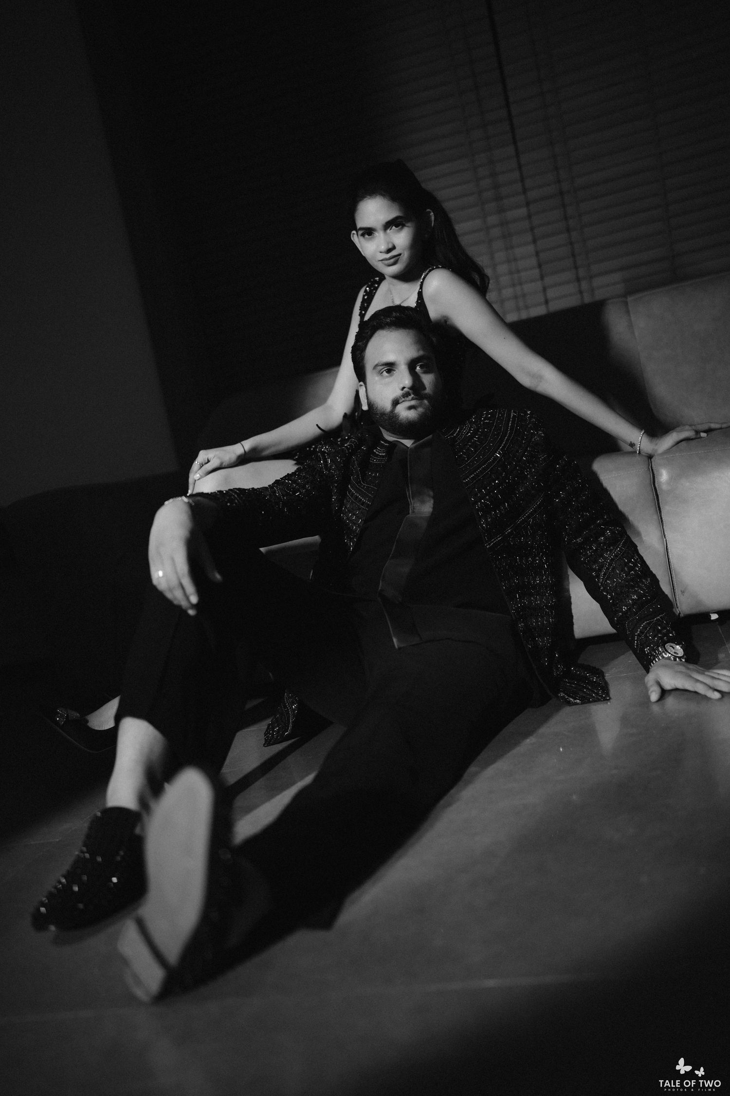 Elegant couple poses in black and white, woman standing behind seated man in stylish dark outfits.