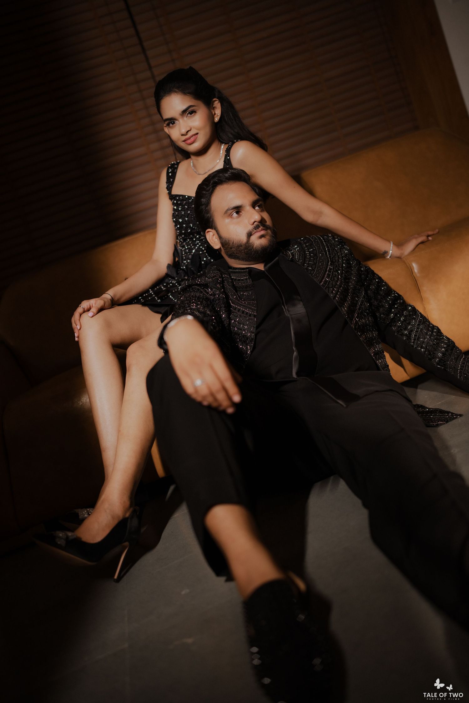 Stylish couple in coordinated black ensembles posing on sofa, woman in sequined dress sitting behind bearded man.