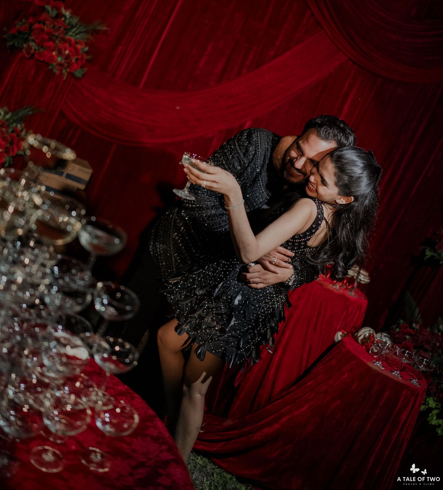 Couple embracing romantically at elegant red-draped event with crystal glassware, both wearing black outfits.