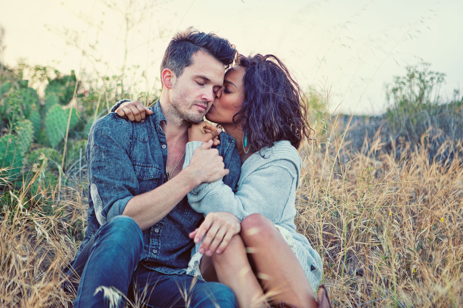 Couple sitting in tall grass sharing a tender moment while holding a small puppy outdoors.