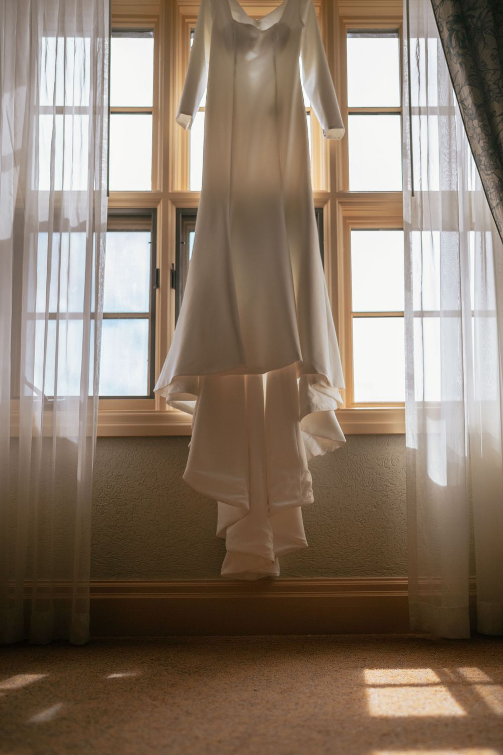 White wedding dress with long sleeves hanging in a sunlit window with sheer curtains.