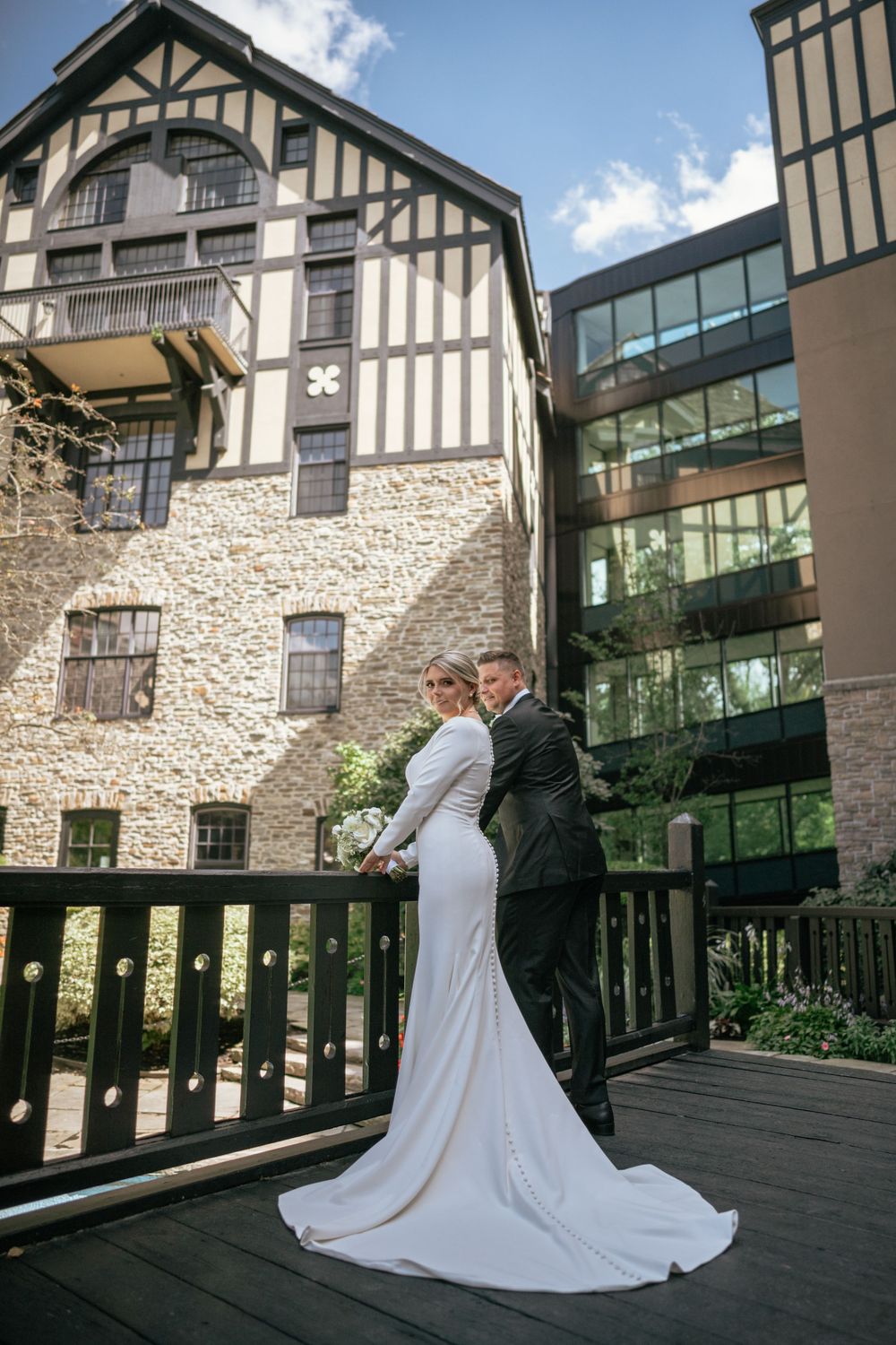 Bride and groom pose on a wooden deck outside a historic stone Tudor building on their wedding day.