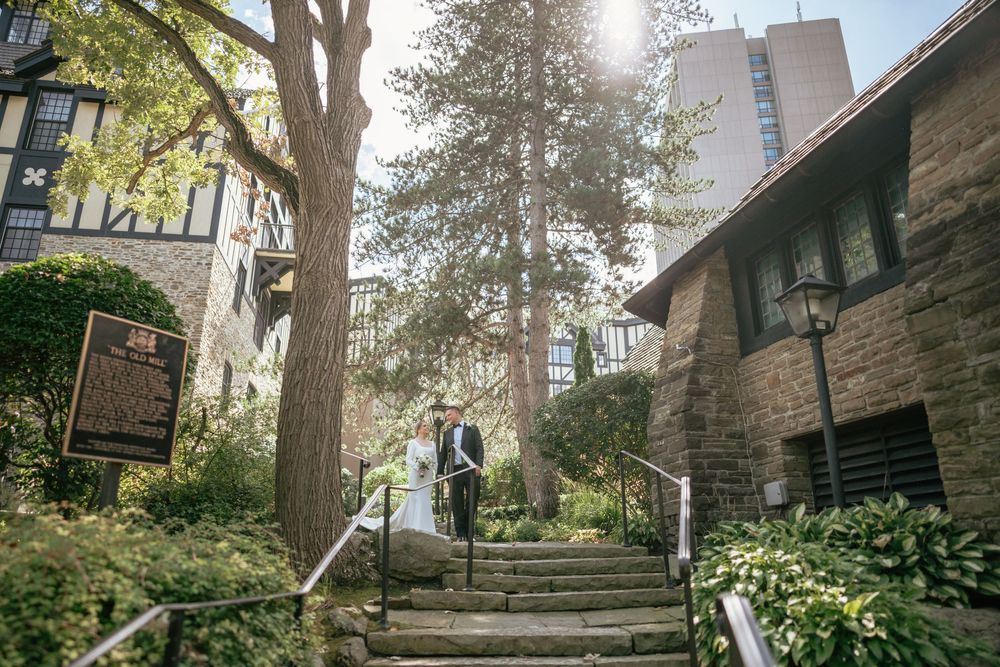 Bride and groom walk down stone steps surrounded by lush greenery and historic buildings on their wedding day.