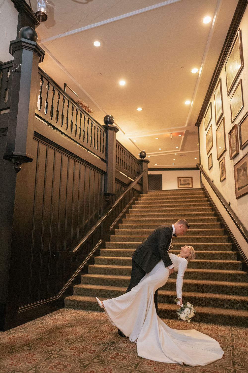 Bride and groom share a romantic dip kiss at the base of an elegant wooden staircase indoors.