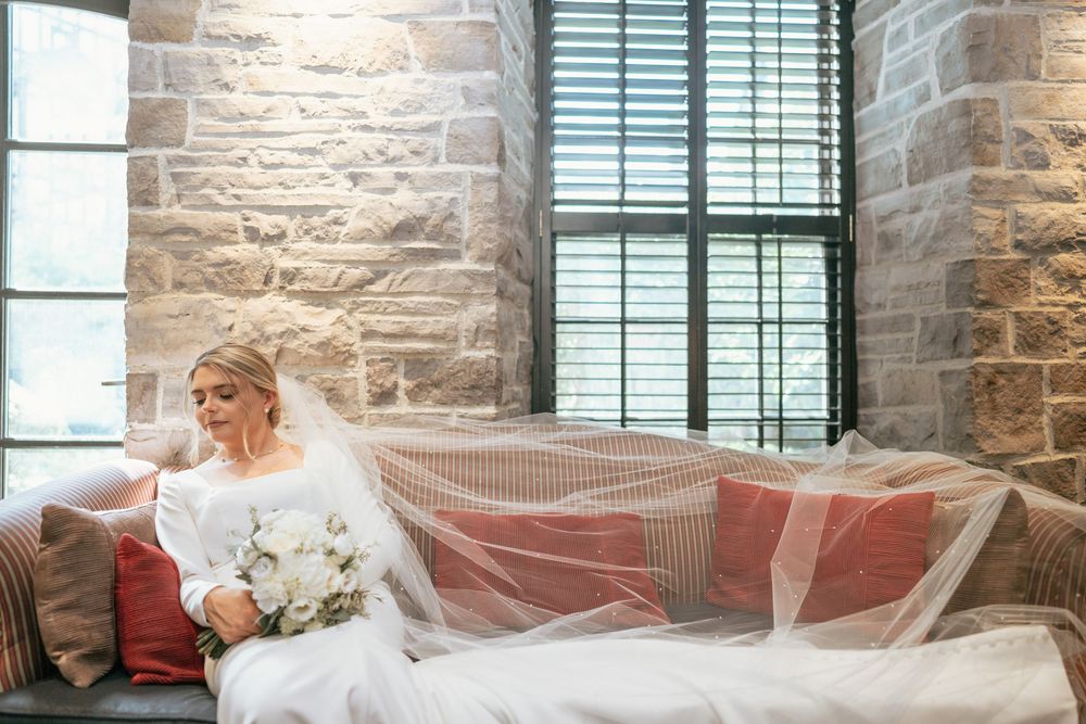 Bride in white gown and veil sits on sofa holding bouquet in stone-walled venue with natural window light.