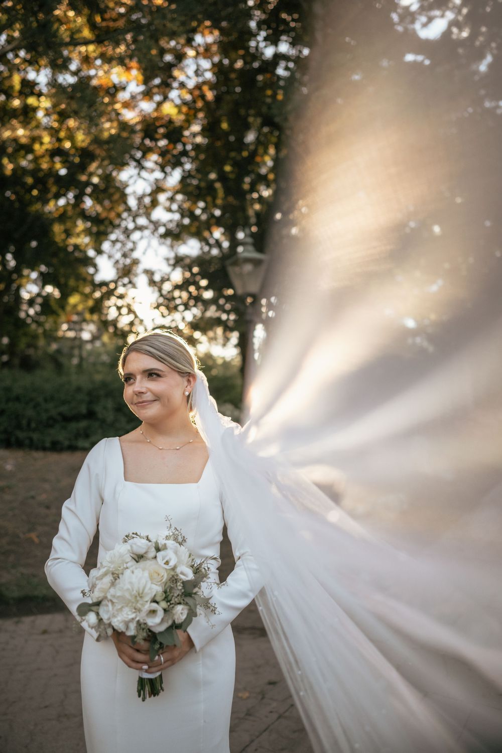 Bride in white gown holds bouquet as veil billows in sunlight, smiling outdoors near trees.