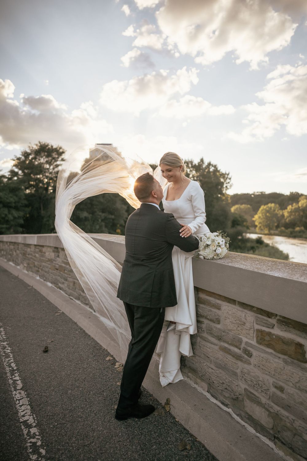 Bride sitting on stone bridge wall, veil blowing in wind, groom in black suit holding her hands, sunset sky backdrop.