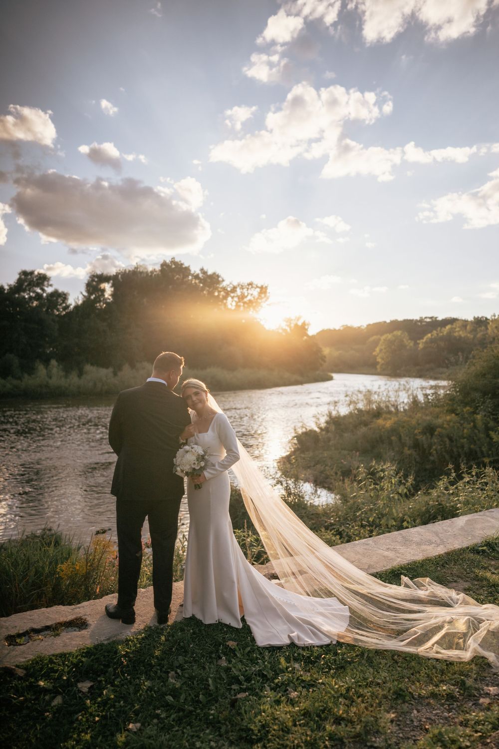 Bride and groom stand together watching a golden sunset over a serene river on their wedding day.