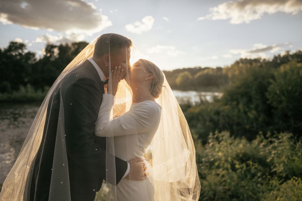 A bride and groom share a romantic kiss under a flowing veil at golden hour by a serene lakeside.