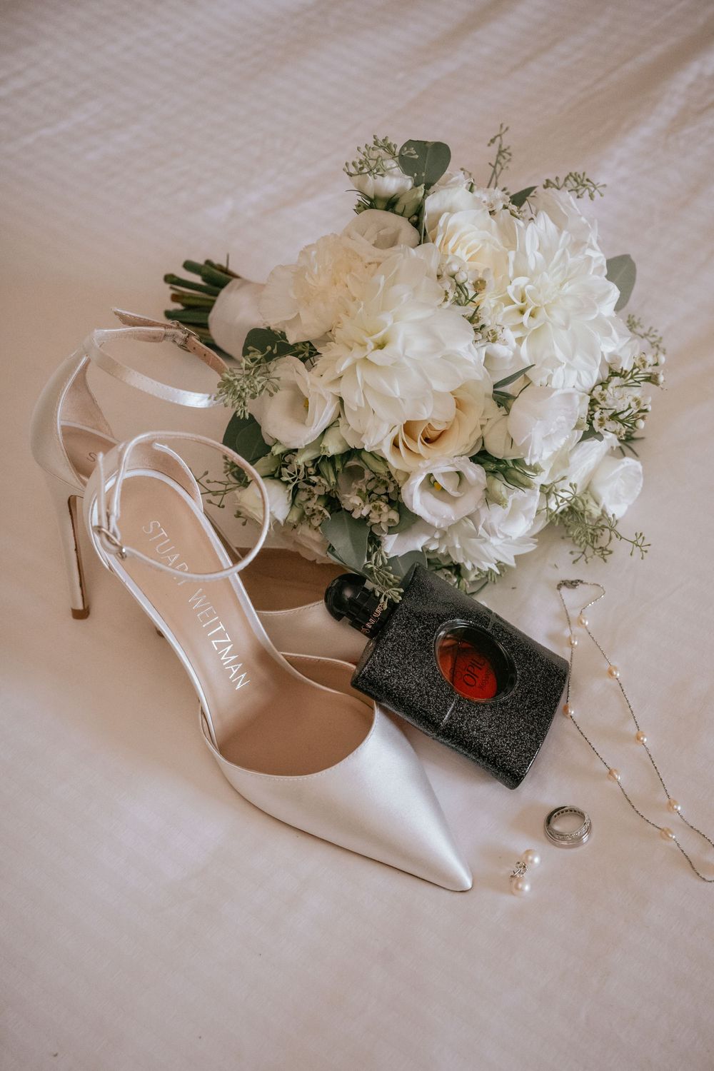 Elegant bridal flat lay featuring Stuart Weitzman heels, YSL Black Opium perfume, white floral bouquet, and delicate jewelry.