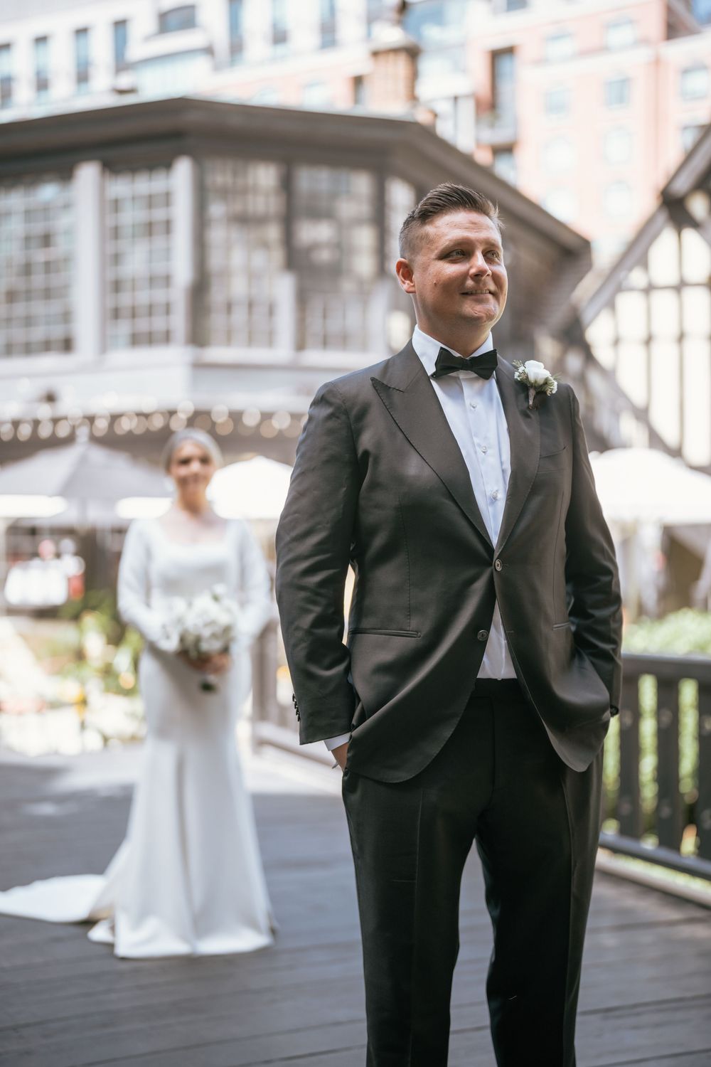 Groom in black tuxedo stands confidently outdoors as bride in white gown approaches for first look.