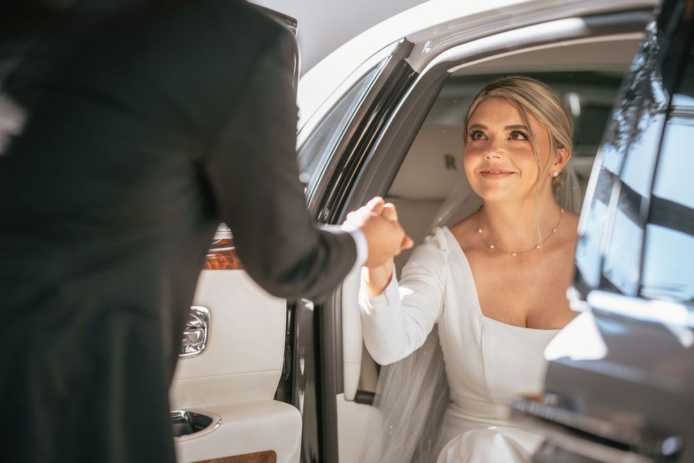 Bride in white gown stepping out of luxury car, assisted by groom in black suit on wedding day.