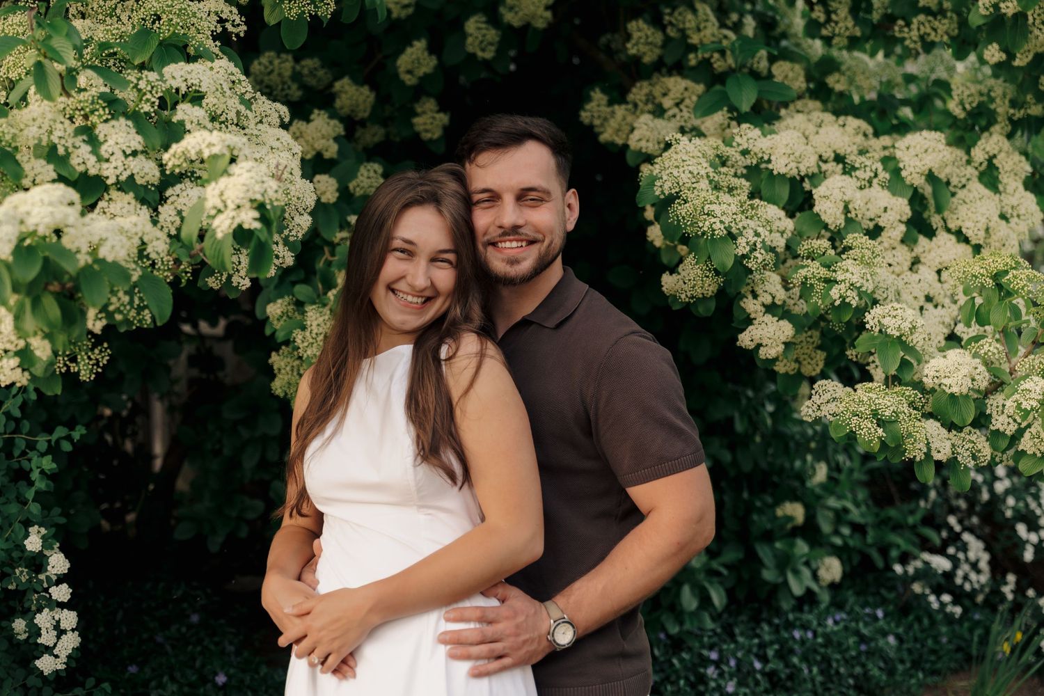 Happy couple embracing outdoors near blooming white flowers, smiling woman in white dress and man in brown polo shirt.