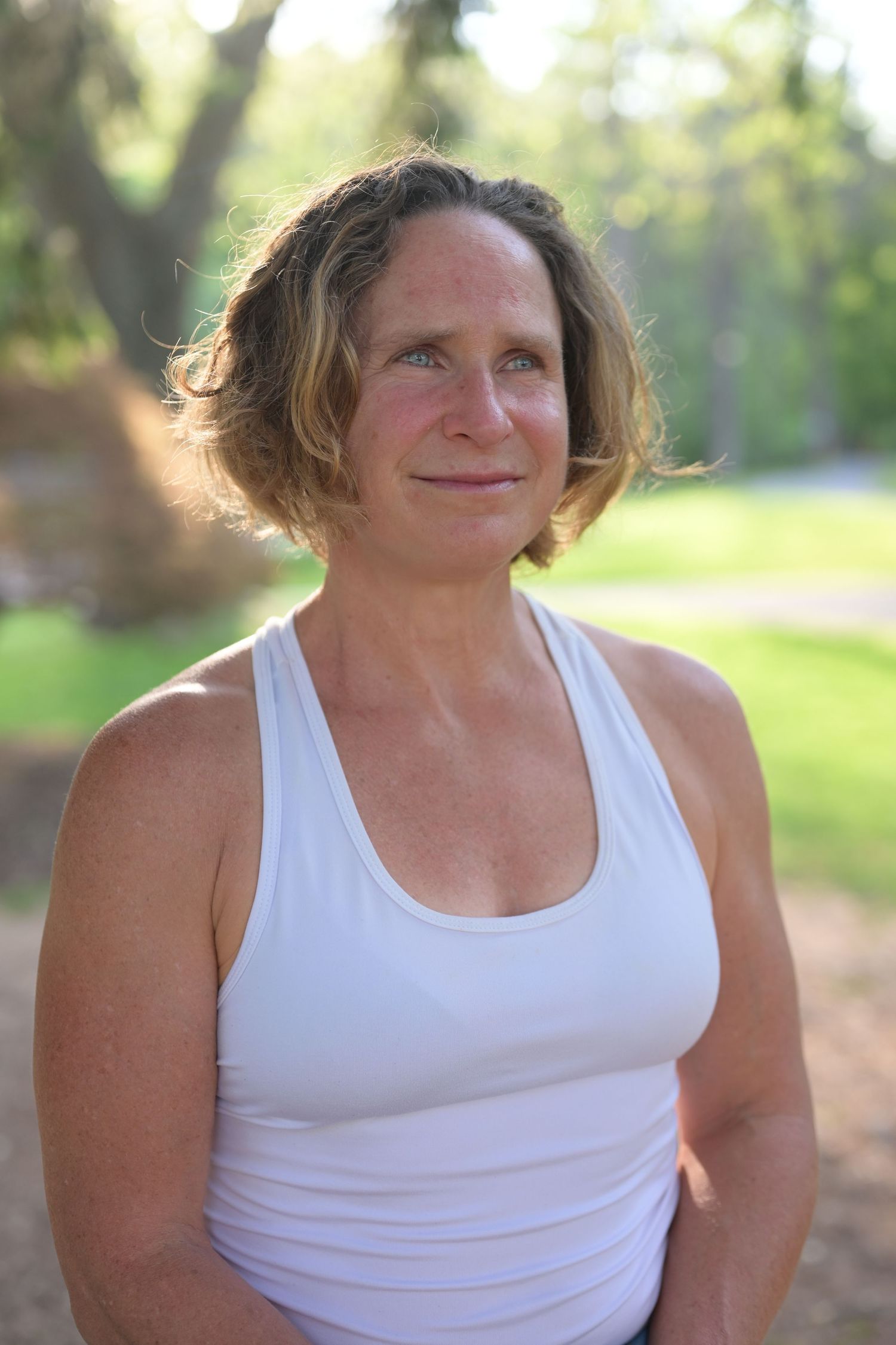Middle-aged woman in white tank top smiling outdoors in a sunny park setting with green trees in background.