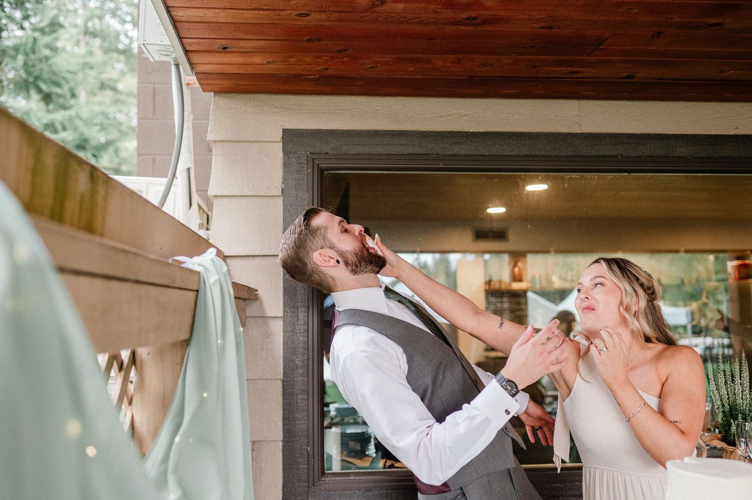 Bride playfully shoves cake in grooms face while laughing together at outdoor backyard wedding