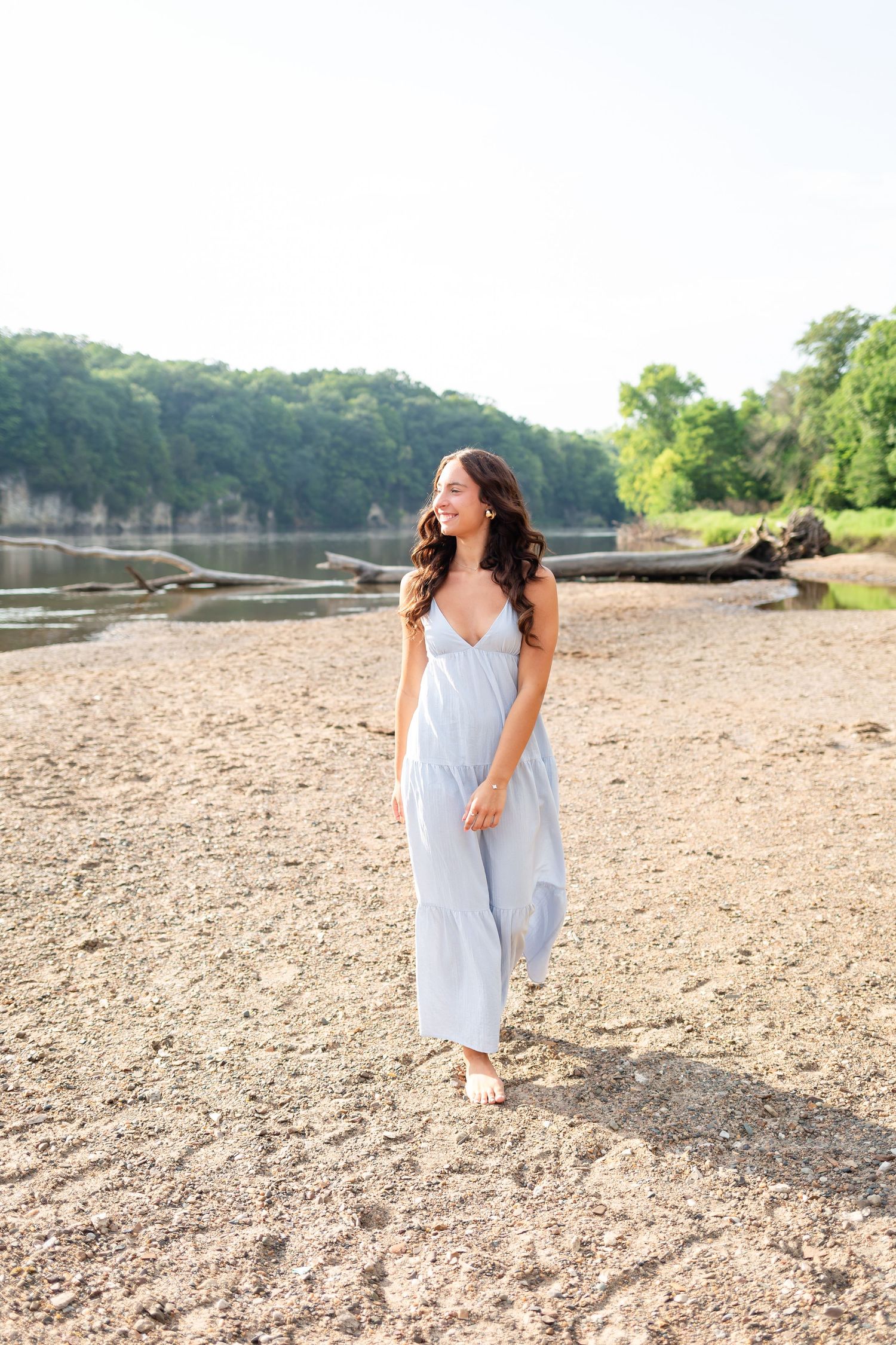 A young woman in a light blue maxi dress walks barefoot on a sandy riverbank surrounded by lush green trees.