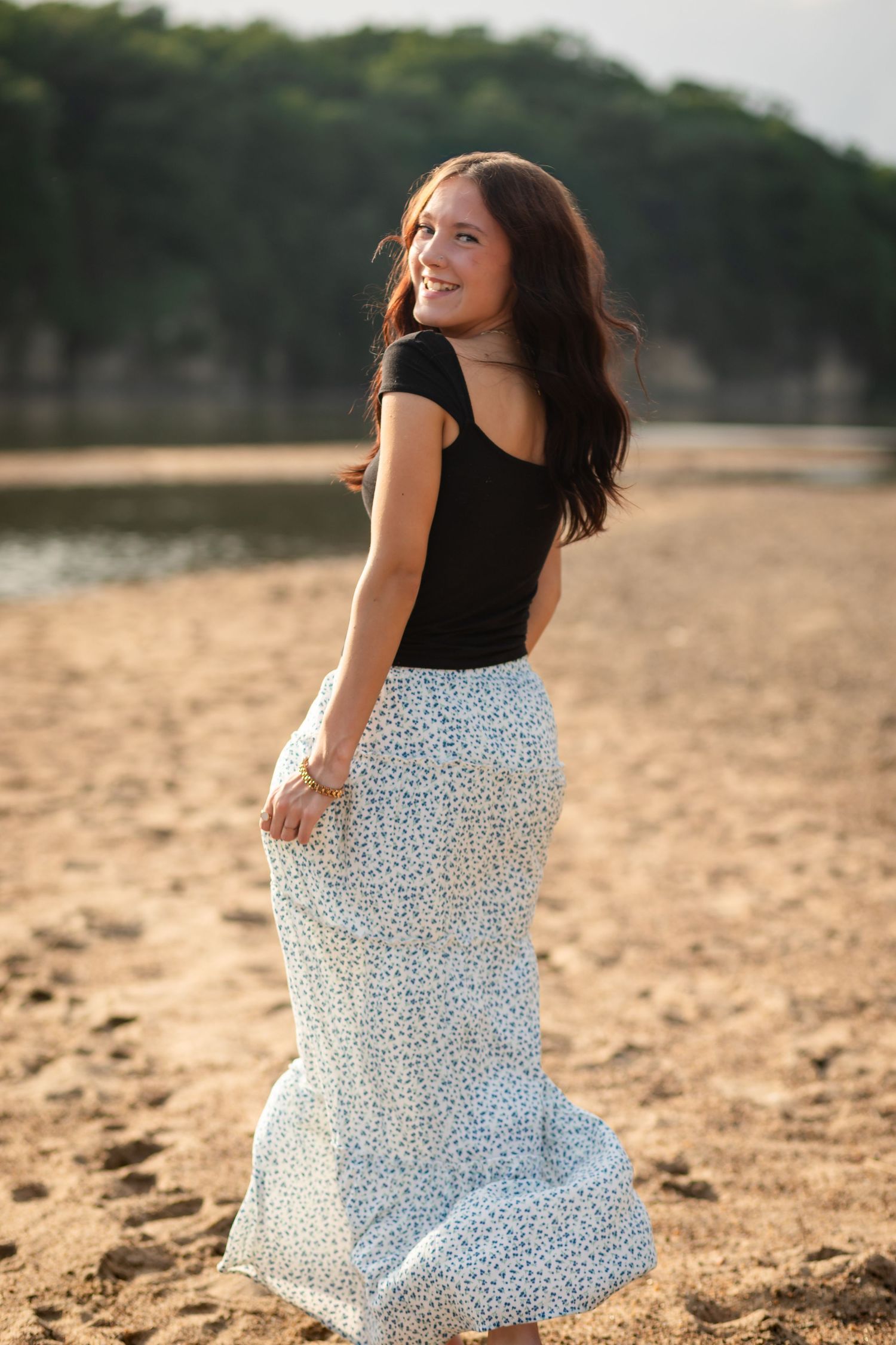 A young woman in a light blue maxi dress walks barefoot on a sandy riverbank surrounded by lush green trees.